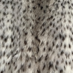 May include: A soft, dense faux fur fabric featuring a light cream and off-white base color with a repeating pattern of small, elongated dark gray and black spots distributed across the entire surface, creating a textured, animal-inspired appearance.