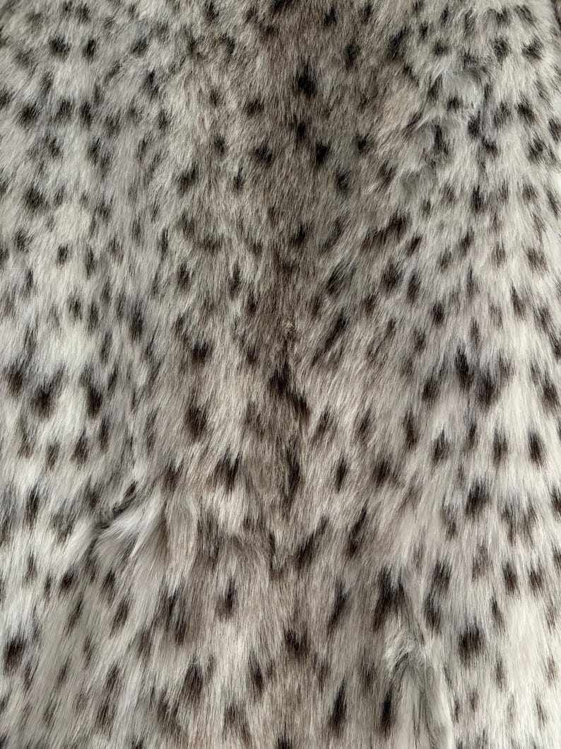 May include: A soft, dense faux fur fabric featuring a light cream and off-white base color with a repeating pattern of small, elongated dark gray and black spots distributed across the entire surface, creating a textured, animal-inspired appearance.