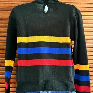 Pu&ograve; includere: Maglione nero con strisce orizzontali gialle, blu e rosse. Il maglione a maniche lunghe ha orlo e polsini a coste. Un piccolo dettaglio a forma di buco della serratura &egrave; visibile sul retro del collo.