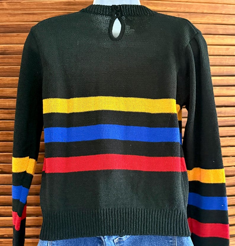Pu&ograve; includere: Maglione nero con strisce orizzontali gialle, blu e rosse. Il maglione a maniche lunghe ha orlo e polsini a coste. Un piccolo dettaglio a forma di buco della serratura &egrave; visibile sul retro del collo.