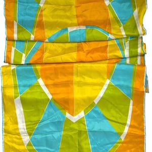 May include: A rectangular scarf featuring a geometric pattern of yellow, orange, lime green, and bright blue segments separated by thin white lines. The fabric has a smooth, slightly reflective finish. The name Una is printed in white in the corner.