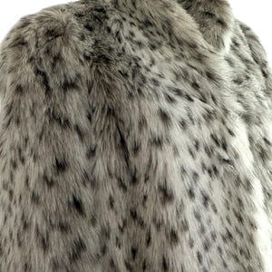 May include: A faux fur coat featuring a light beige and grey base color with a dense pattern of dark brown and black spots throughout. The garment has a high collar and a thick, textured pile that mimics the appearance of animal fur.