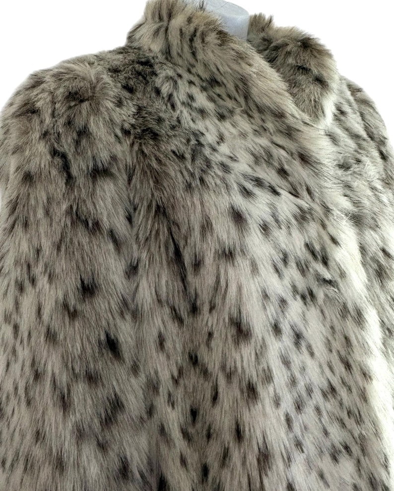 May include: A faux fur coat featuring a light beige and grey base color with a dense pattern of dark brown and black spots throughout. The garment has a high collar and a thick, textured pile that mimics the appearance of animal fur.