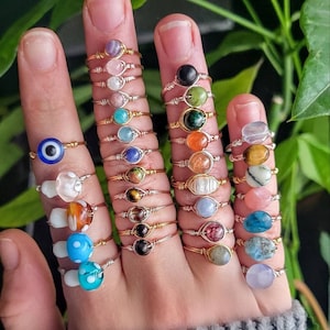 May include: A collection of wire-wrapped rings featuring various colourful gemstones, glass beads, and an evil eye charm. The rings are worn on fingers, showcasing diverse textures, shapes, and hues including blue, pink, green, orange, and clear stones.