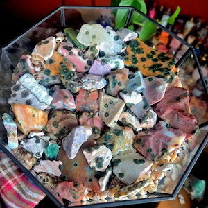Puede incluir: Un terrario de vidrio lleno de una variedad de rocas coloridas y conchas marinas. Las rocas muestran una gama de colores, incluyendo naranja, rosa, verde y blanco, con patrones únicos. El terrario tiene una forma geométrica.
