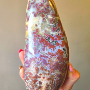 Puede incluir: Una piedra de jaspe oceánico pulida y alargada que presenta un patrón complejo de orbes circulares en tonos rojo intenso, granate y lavanda suave, sobre un fondo translúcido de color blanco lechoso y gris con inclusiones minerales naturales.