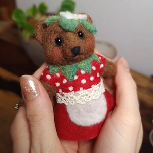 Puede incluir: Un oso de peluche marrón hecho a mano que lleva un disfraz de fresa rojo con lunares blancos, un cuello verde y un delantal de encaje blanco. El oso tiene ojos negros y una flor blanca en la cabeza. Es una artesanía de fieltro.