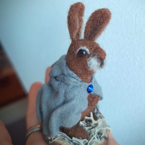 May include: A handcrafted felted rabbit figurine. The brown rabbit wears a gray hooded cloak with a blue gem clasp. The rabbit is perched on a small, textured base, with a light blue background.