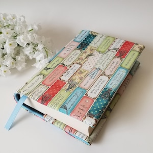 May include: A fabric book cover with a colorful design featuring book-themed text and illustrations. The cover has a light blue ribbon bookmark. The text includes phrases like "Once upon a time" and "My weekend is all booked!"