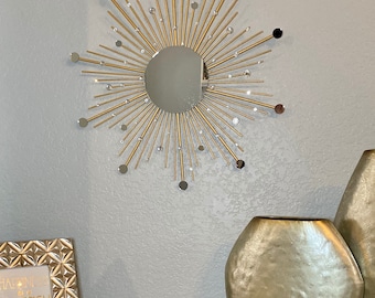 May include: A gold sunburst mirror with a round mirror in the center. The mirror is surrounded by gold metal rays with small clear rhinestones. The mirror is hanging on a light gray wall.