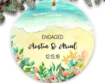 May include: A round, white ceramic ornament with a watercolor beach scene. The ornament features a blue ocean, white waves, and a yellow sandy beach. The text "ENGAGED Austin & Arial 12.15.18" is written on the ornament.