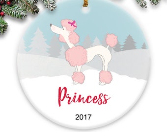 Könnte beinhalten: Weißer Porzellan-Weihnachtsbaumschmuck mit einer Cartoon-Illustration eines rosa Pudels mit einer rosa Schleife. Der Anhänger hat den Text "Princess" und "2017" aufgedruckt.