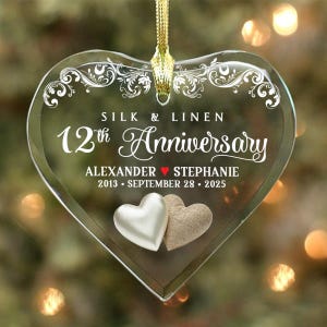 May include: Clear heart-shaped ornament with gold ribbon. The ornament has the text "SILK & LINEN 12th ANNIVERSARY" and the names "ALEXANDER ❤️ STEPHANIE" with the date "2013 • SEPTEMBER 28 • 2025". Two heart-shaped embellishments are at the bottom.