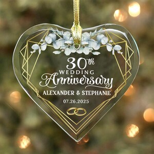 May include: Heart-shaped clear glass ornament with gold geometric design and text. The ornament reads "30th Wedding Anniversary" with names and a date. A gold ribbon is attached for hanging. Eucalyptus sprigs are at the top.