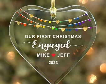 May include: A clear glass heart-shaped Christmas ornament with a rainbow of lights and the text "Our First Christmas Engaged Mike ♥ Jeff 2023"