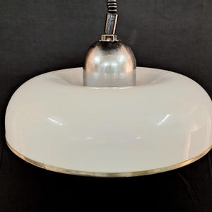 May include: A white, donut-shaped pendant light with a polished metal top and a gold-colored trim. The light fixture hangs from a flexible, black cord. The design is modern and minimalist, suitable for various interior styles.