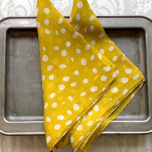 May include: Two yellow polka dot napkins are neatly folded on a vintage silver tray with ornate handles. The napkins have a repeating pattern of white polka dots. The tray sits on a white patterned surface.