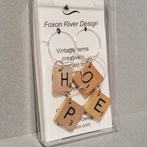 May include: A set of four wine glass charms featuring the word "HOPE" spelled out using wooden Scrabble tiles. The charms are packaged in a clear plastic box with a white card that reads "Foxon River Design".