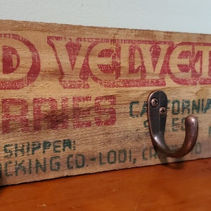 May include: A wooden sign with the text "RED VELVET CHERRIES CALIFORNIA" and three hooks. The sign is painted in red, green, and white. The hooks are made of metal and have a brown finish.