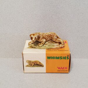 Może przedstawiać: Ceramiczna figurka leoparda w kolorze brązowym i beżowym, siedzącego na skale. Figurka znajduje się w pudełku z napisem "Whimssies" i "Wade Made in England".