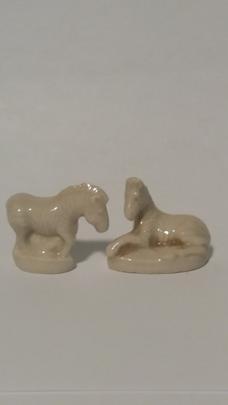 Może przedstawiać: Dwie ceramiczne figurki zebr, jedna stojąca, a druga leżąca. Obie są jasnobeżowe.