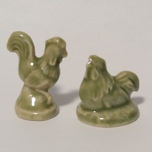 Może przedstawiać: Dwie zielone ceramiczne figurki kogut&oacute;w. Jeden kogut stoi z uniesionymi pi&oacute;rami ogona. Drugi kogut siedzi z głową odchyloną do tyłu.