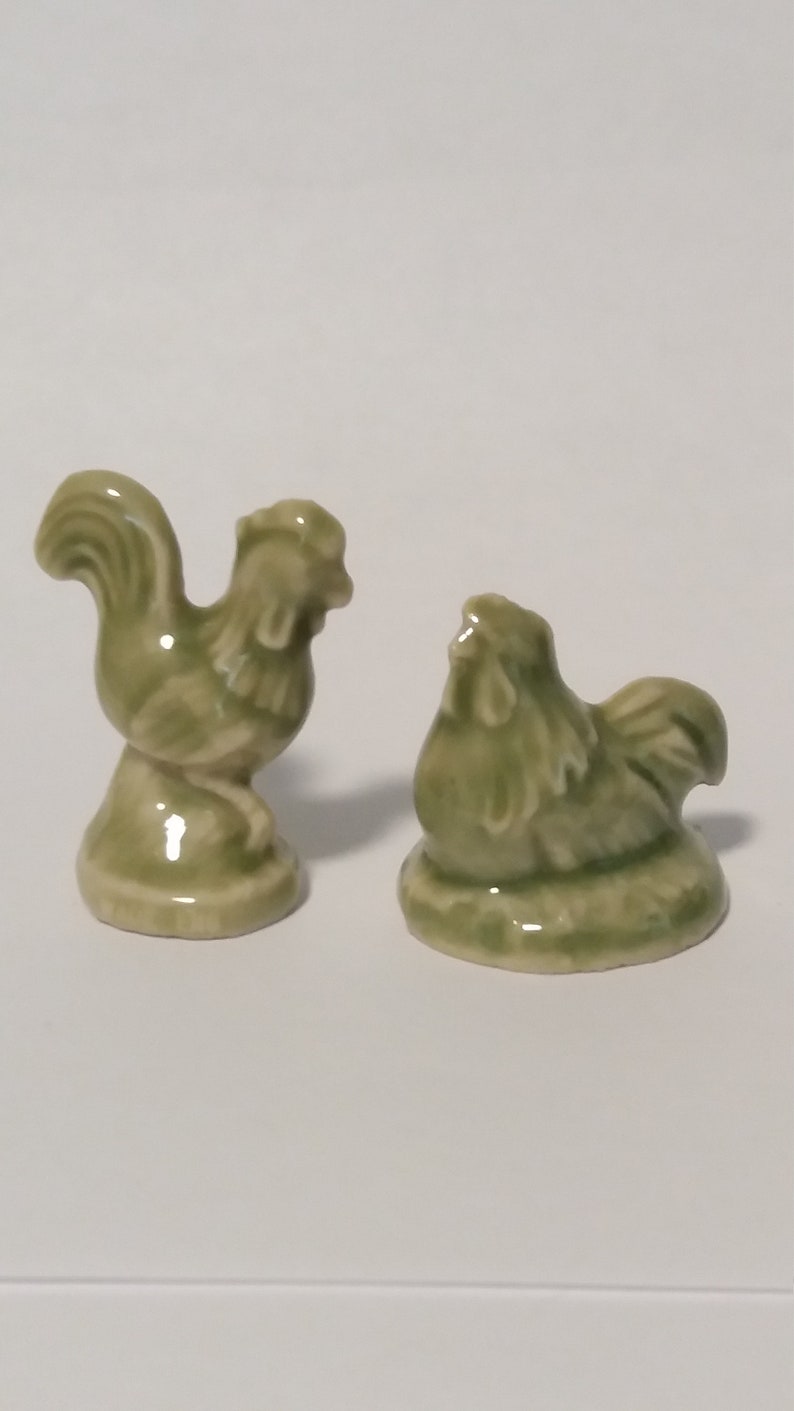 Może przedstawiać: Dwie zielone ceramiczne figurki kogut&oacute;w. Jeden kogut stoi z uniesionymi pi&oacute;rami ogona. Drugi kogut siedzi z głową odchyloną do tyłu.