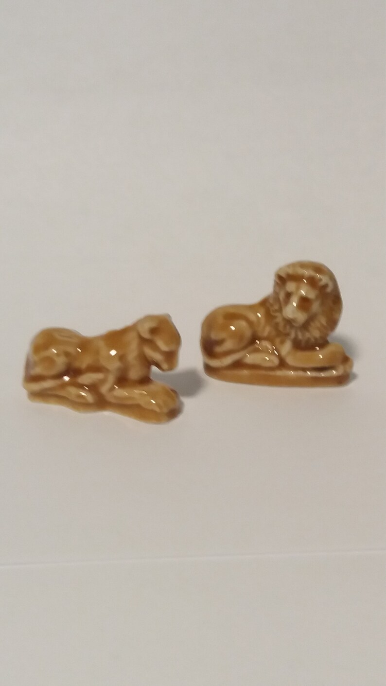 Może przedstawiać: Dwie ceramiczne figurki lw&oacute;w, jedna leżąca, a druga siedząca. Obie są jasnobrązowe.