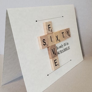 May include: A white card with a cross made of wooden Scrabble tiles spelling out "SIXTY". The tiles are arranged so that the "V" in "SIXTY" is on top of the "E". Below the tiles is the text "Is only 25 in SCRABBLE".