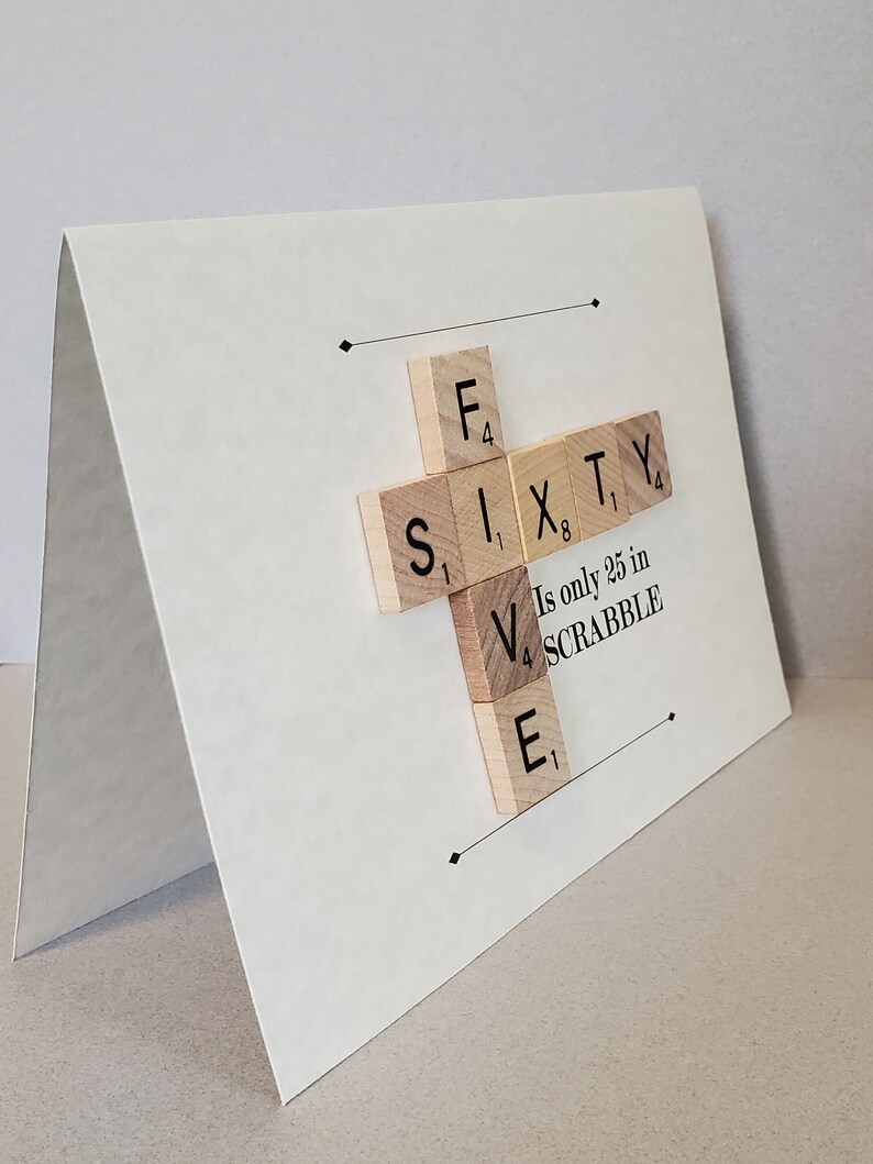 May include: A white card with a cross made of wooden Scrabble tiles spelling out "SIXTY". The tiles are arranged so that the "V" in "SIXTY" is on top of the "E". Below the tiles is the text "Is only 25 in SCRABBLE".