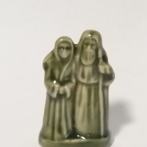 Może przedstawiać: Zielona ceramiczna figurka przedstawiająca parę stojącą razem. Mężczyzna trzyma laskę, a kobieta trzyma ręce przed sobą.