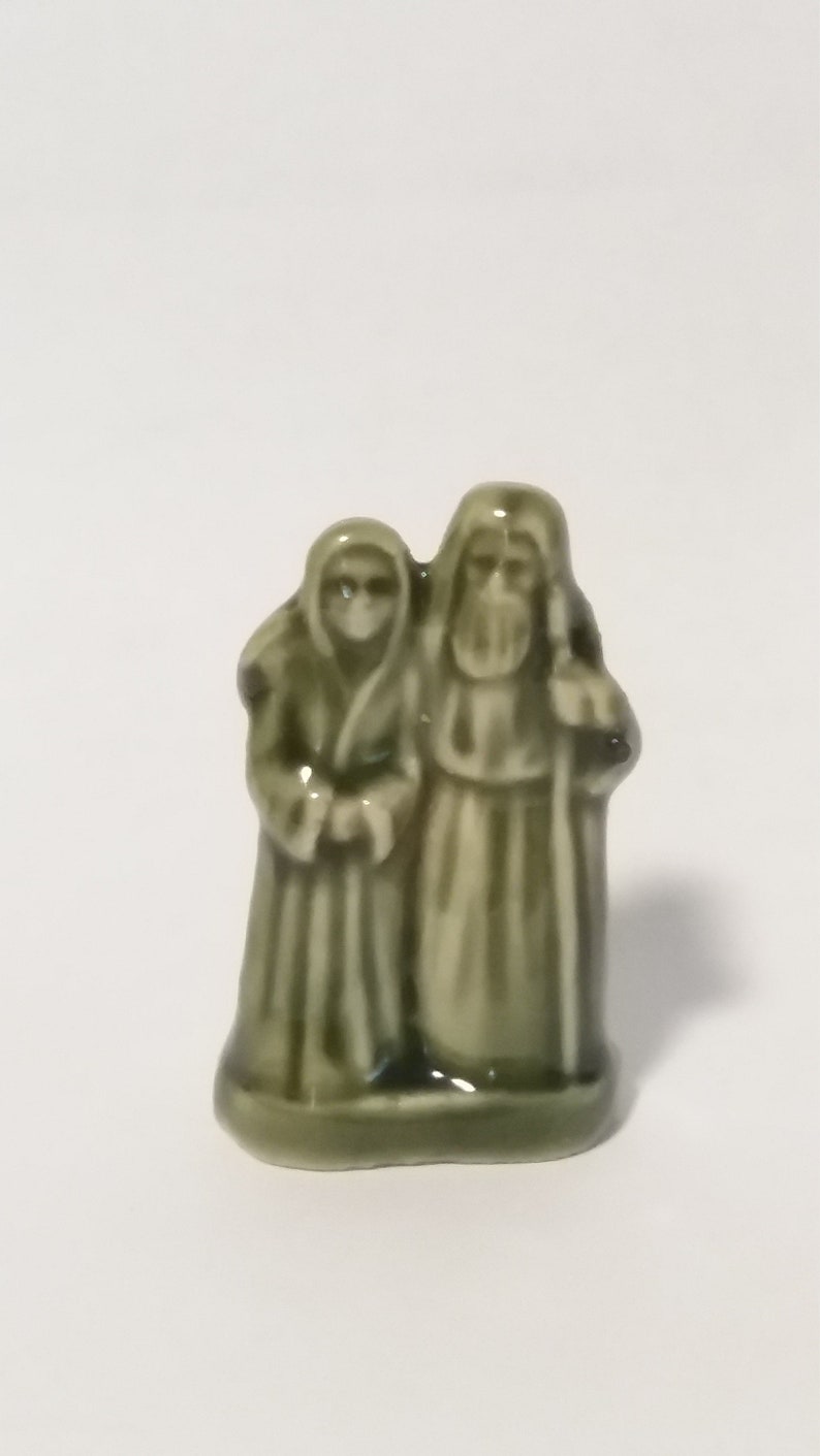 Może przedstawiać: Zielona ceramiczna figurka przedstawiająca parę stojącą razem. Mężczyzna trzyma laskę, a kobieta trzyma ręce przed sobą.
