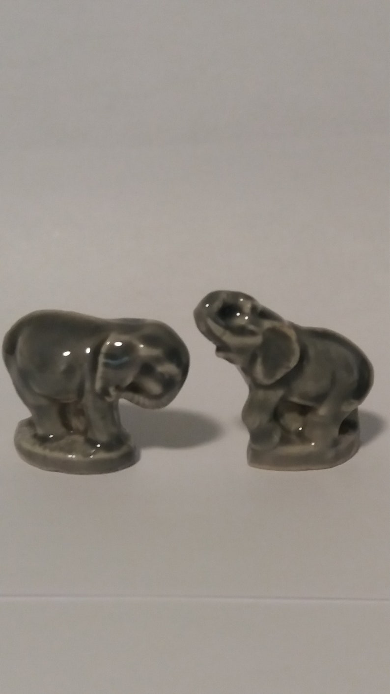 Może przedstawiać: Dwie szare ceramiczne figurki słoni. Jeden słoń stoi z uniesioną trąbą, podczas gdy drugi siedzi z zwiniętą trąbą.