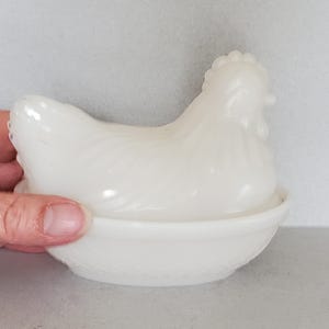 May include: A white ceramic hen-shaped egg holder. The hen sits atop a basket-like base with a textured pattern. The lid of the hen is removable, revealing the space for eggs. The piece is a vintage-style kitchen decor item.