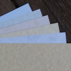 May include: A stack of six different colored paper sheets, ranging from light blue to beige, with a textured surface. The paper is arranged in a fan shape, with the lightest color on top and the darkest color on the bottom.