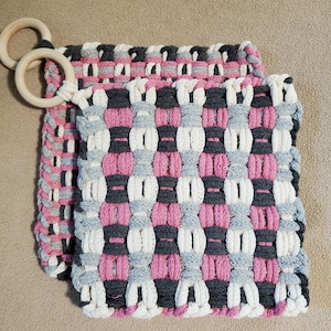 May include: Two woven pot holders with a pink, gray, and white checkered pattern. The pot holders have a wooden ring attached to the top.