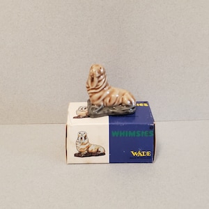Może przedstawiać: Mała ceramiczna figurka morsa siedzącego na skale. Figurka jest brązowa i biała i ma pudełko z napisem "Whimssies" i "Wade".