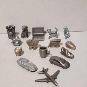 May include: A selection of small, silver-coloured metal figurines. The figurines include a dog, cat, penguin, laptop, car, airplane, shoe, and other objects. The objects are arranged on a light grey surface.
