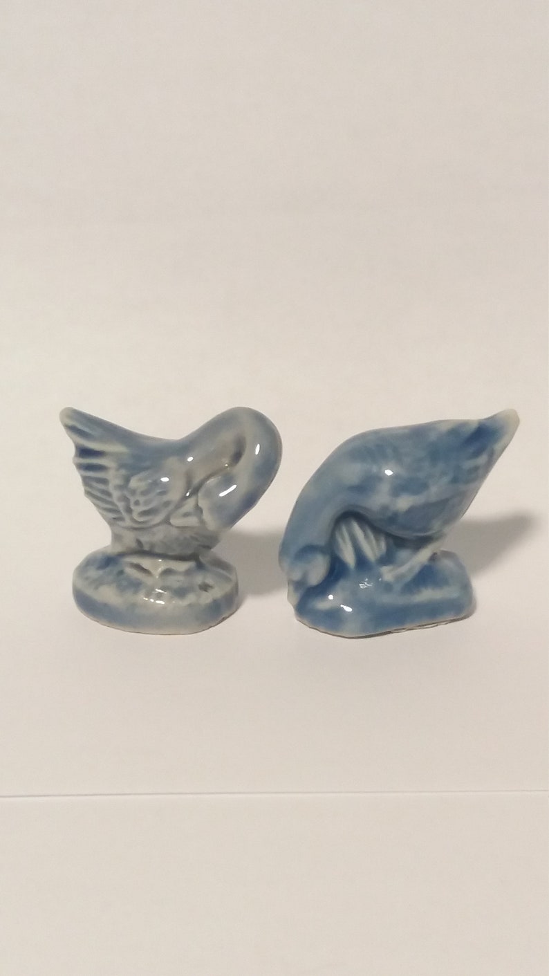 Może przedstawiać: Dwie niebieskie ceramiczne figurki gęsi. Jedna gęś stoi z głową opuszczoną, podczas gdy druga gęś stoi z głową uniesioną.