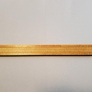 May include: A vintage wooden slide rule with etched markings and numbers. The slide rule is a light brown color and has a rectangular shape. The markings are in black and red. The slide rule is a tool used for mathematical calculations.