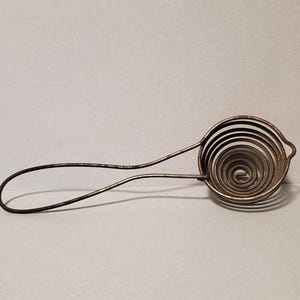 May include: A vintage egg separator made of metal. The tool features a spiral-shaped wire head and a long handle with a loop at the end. The metal has a tarnished appearance, suggesting age and use. The design is simple and functional.