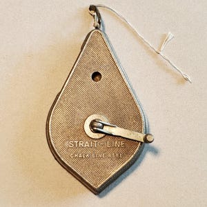 May include: A vintage silver-toned chalk line reel with a diamond-shaped body. The reel has a textured surface and a crank for winding the line. The text "STRAIT-LINE CHALK LINE REEL" is printed on the reel.
