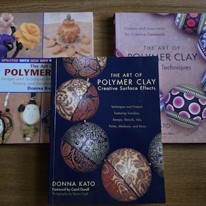 May include: Three instructional books on polymer clay techniques by Donna Kato. The books feature colorful illustrations of clay projects, including floral designs, sculpted faces, and patterned spheres, with titles like The Art of Polymer Clay.