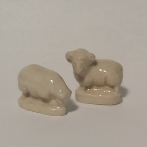 Może przedstawiać: Dwie ceramiczne figurki owiec. Jedna owca stoi z podniesioną głową, podczas gdy druga leży z opuszczoną głową. Obie owce są białe i mają prosty design.