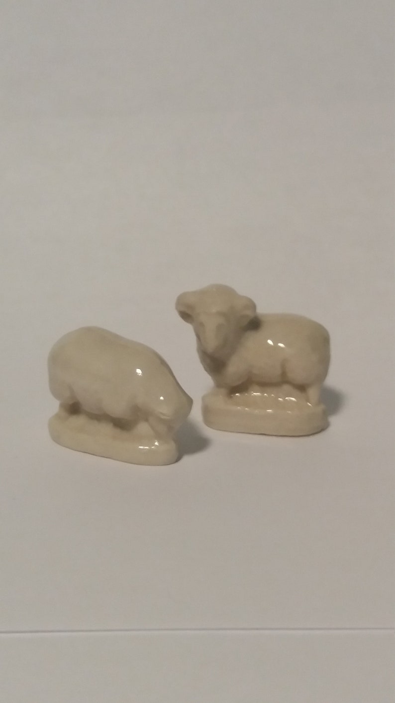 Może przedstawiać: Dwie ceramiczne figurki owiec. Jedna owca stoi z podniesioną głową, podczas gdy druga leży z opuszczoną głową. Obie owce są białe i mają prosty design.