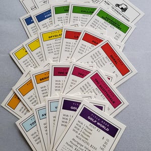 May include: A collection of Monopoly Title Deed cards, featuring various golf course properties. The cards have colorful borders and text detailing rent costs and property values. The cards are scattered on a light gray surface.