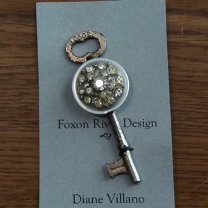 May include: A key-shaped brooch with a clear, round centrepiece set with rhinestones. The brooch has a silver-coloured stem and key details. The card behind it reads "Foxon Riv Design" and "Diane Villano".