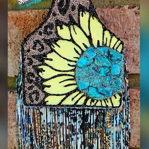 May include: A brown and black leather tag with a yellow sunflower and turquoise gemstone accents. The tag has a fringe of colorful fabric with a floral pattern.
