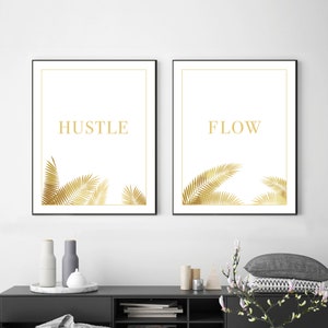 May include: Two framed art prints with black frames. The prints feature the words "HUSTLE" and "FLOW" in gold lettering, with gold palm fronds at the bottom. The background is white. The prints are displayed above a black cabinet.