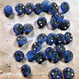 Puede incluir: Una colección de botones de vidrio azul con diseños florales en blanco y dorado. Los botones están esparcidos sobre un fondo de papel marrón.